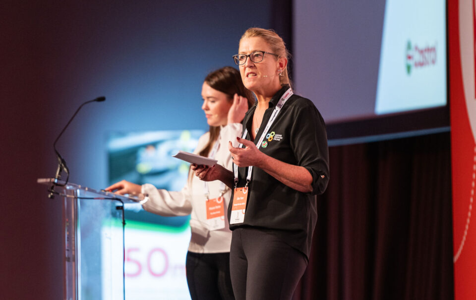 A speaker on stage at The Blend 2026 conference, presenting to an audience of UK independent garage owners.