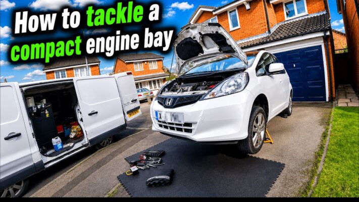 A mechanic performs a Honda Jazz clutch replacement, with the car raised on axle stands and the engine bay visible.