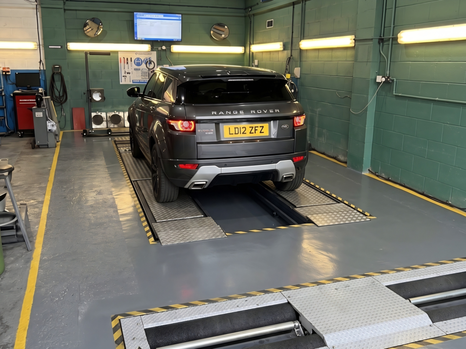 A DVSA enforcement officer observing one of the UK's MOT testers conducting a vehicle inspection in a garage workshop.
